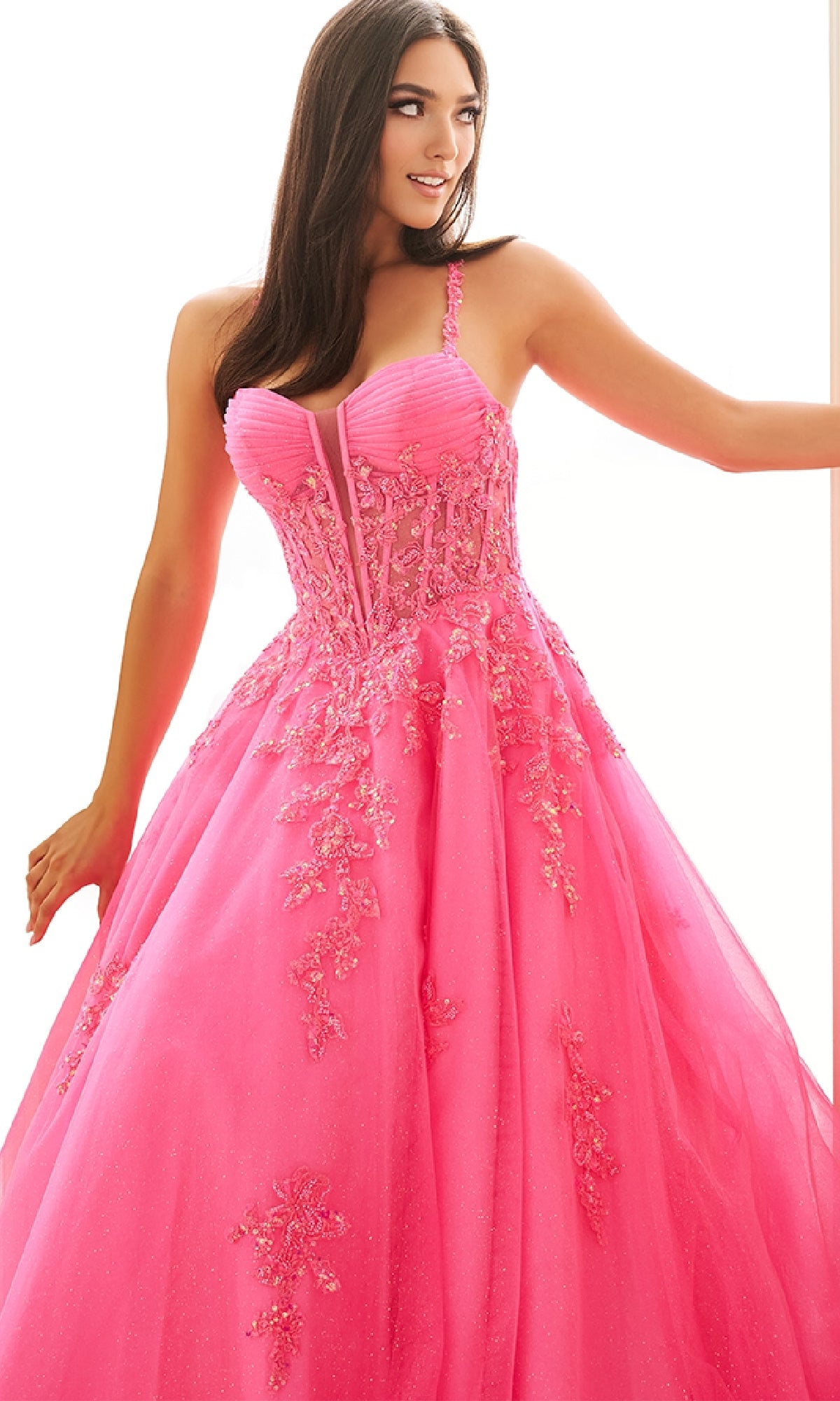 A woman wears the Long Prom Dress: Ellie Wilde EW36202, a bright pink ball gown with a corset bodice, floral embroidery, and fitted silhouette, standing against a white background.